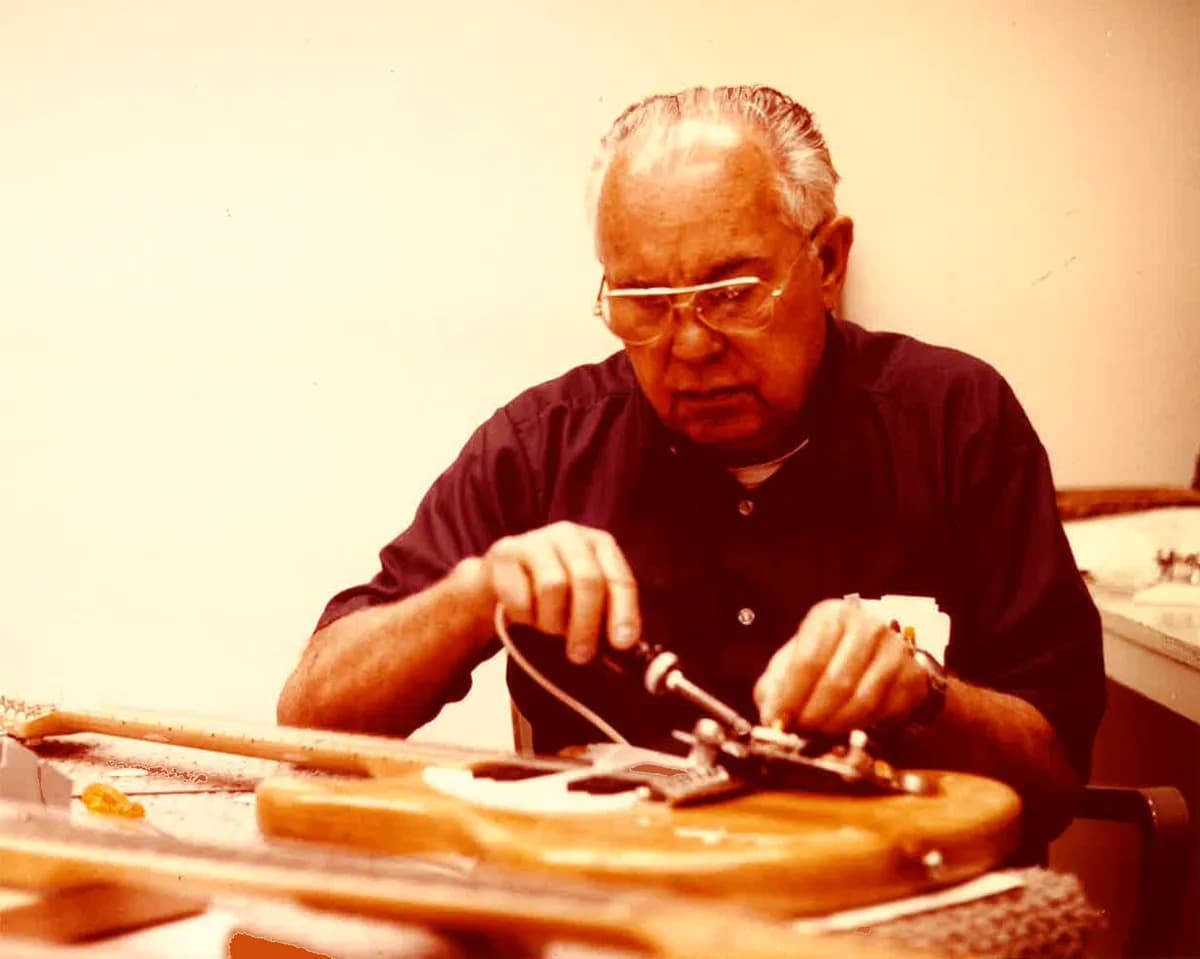 Leo Fender, in glasses and a dark shirt, soldering the electronics into a Telecaster-style guitar body at his workbench.